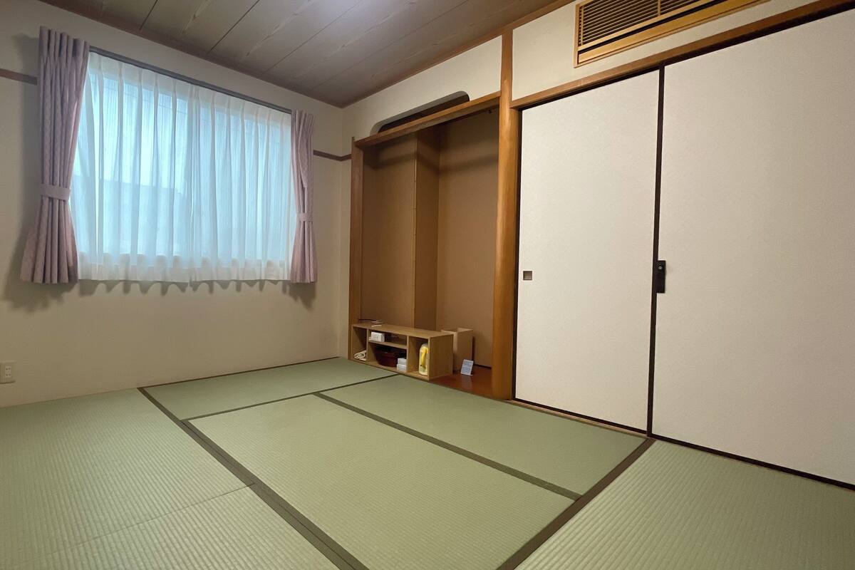 Una habitación con suelo de tatami, puerta corredera, ventana con cortinas y estante con objetos.