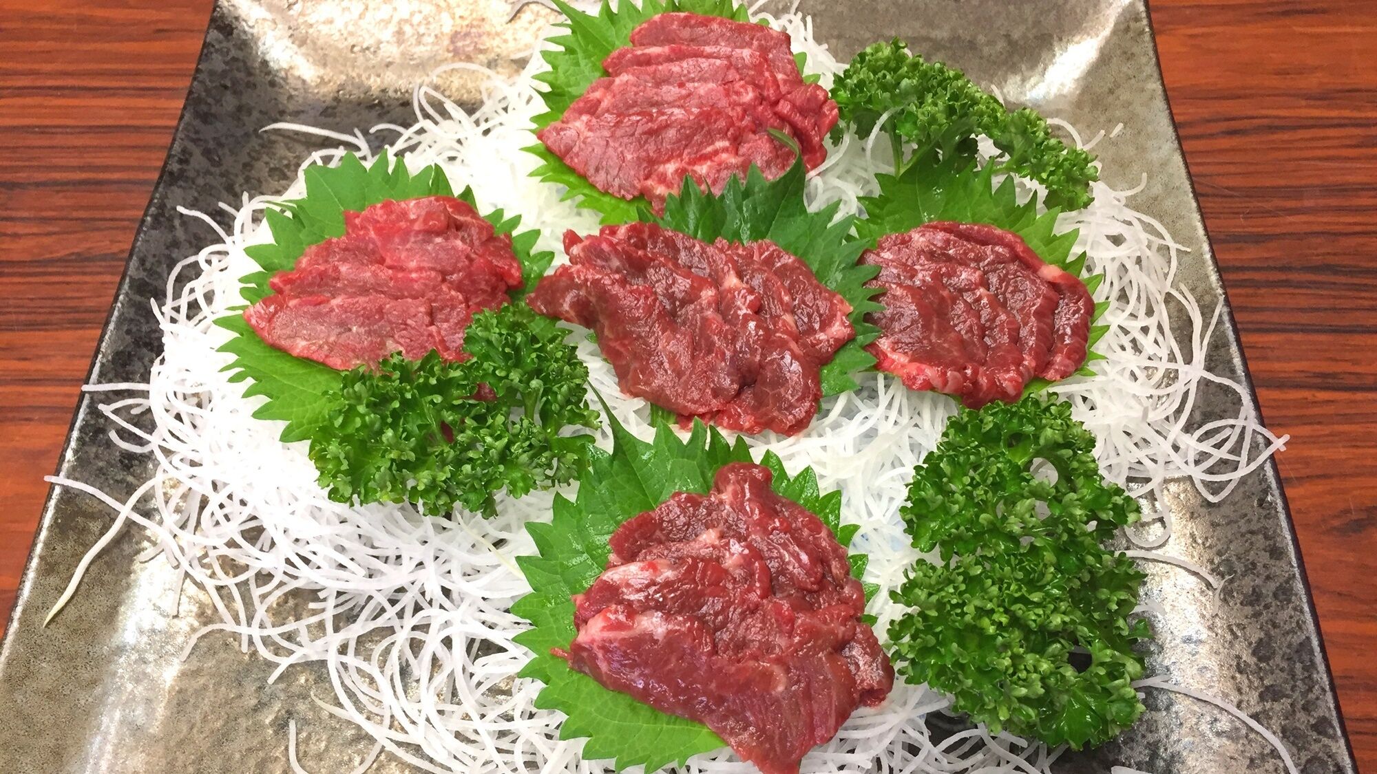 A tray with raw beef and green leaves on a metal surface.
