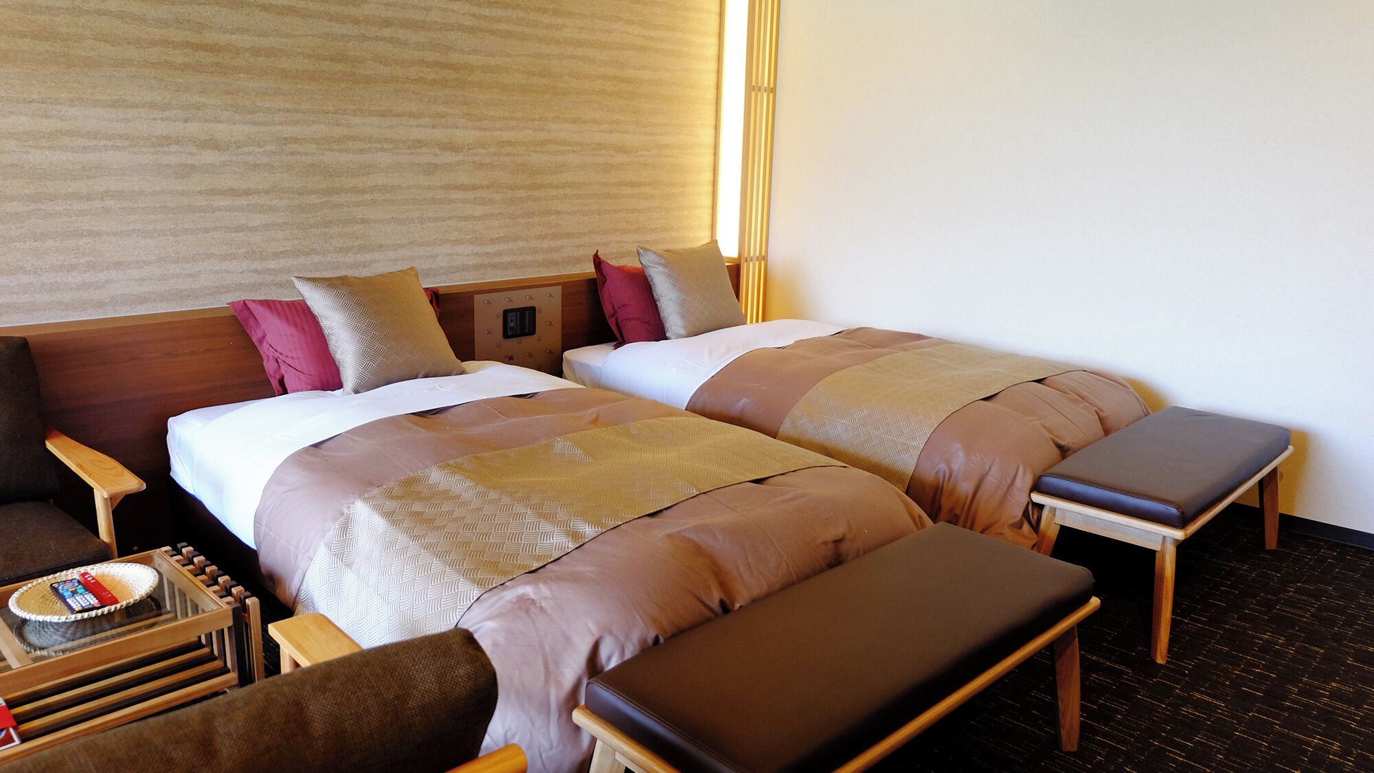 A hotel room with two beds, a wooden nightstand, a small table with a phone, and a bench.
