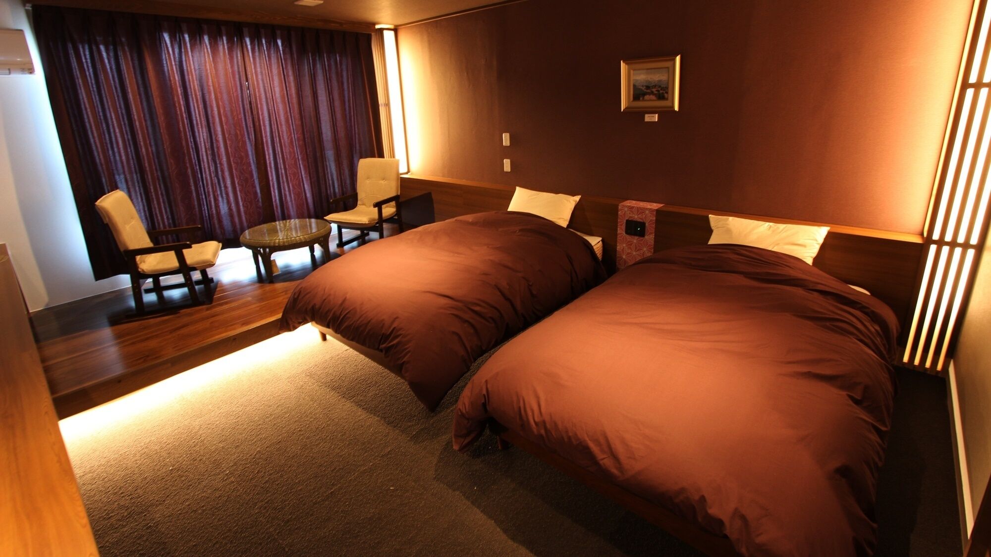 Une chambre d’hôtel avec deux lits, une petite table, des chaises et une grande fenêtre avec des rideaux.