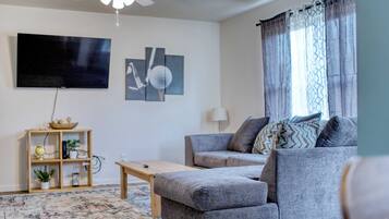 Un salon avec un canapé gris, une table basse en bois, un téléviseur fixé au mur et un ventilateur de plafond.