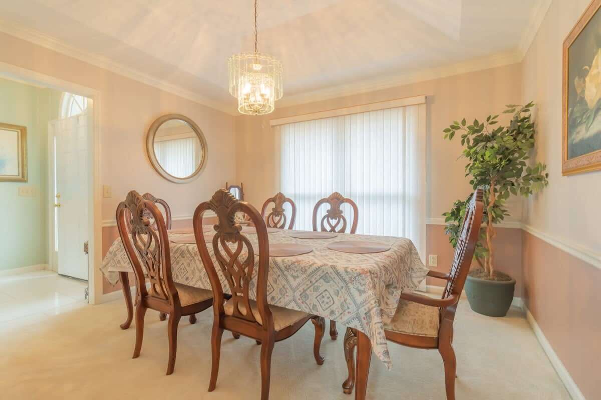 Une salle à manger avec une table dressée pour quatre personnes, entourée de chaises en bois, d’un lustre, d’un grand miroir et d’une plante en pot.