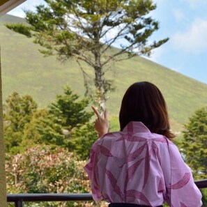 A person standing on a balcony with a view of a hill and trees. - Izukogen Hanairotei (Ito)