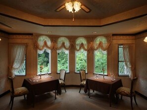 A dining room with a high ceiling, chandeliers, and a view of greenery through large windows. - Izukogen Hanairotei (Ito)