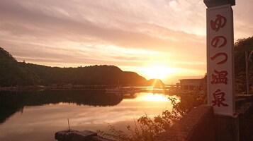 Un sereno tramonto su un lago calmo, con una barca ormeggiata sulla riva, un cartello con caratteri giapponesi e un paesaggio collinare sullo sfondo.