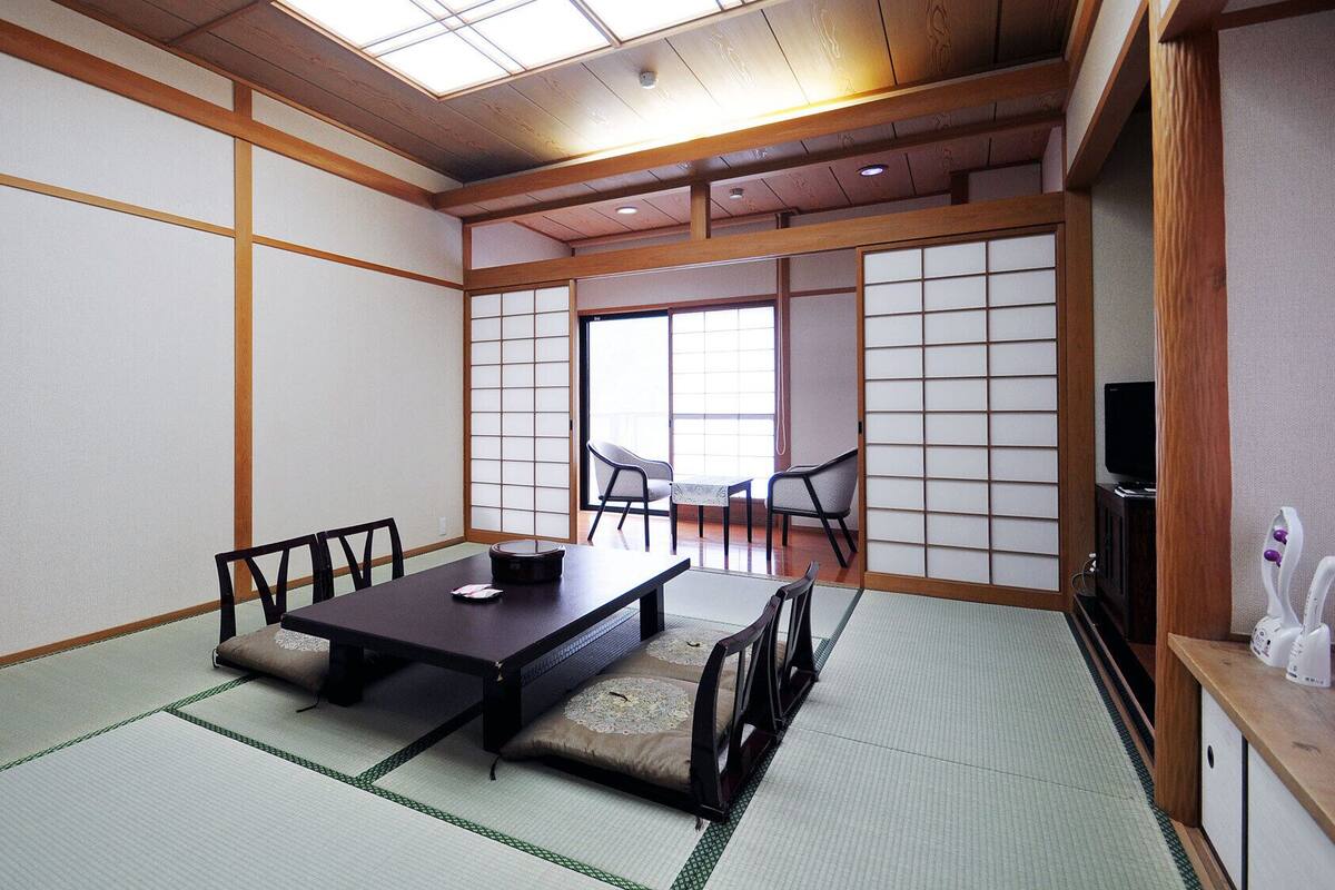 A traditional Japanese room with tatami flooring, a low table, chairs, sliding doors, and a television.