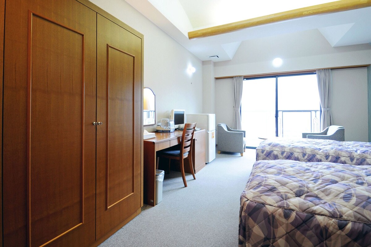 A hotel room with a large wardrobe, a desk with a computer, a chair, a bed, and a window with curtains.