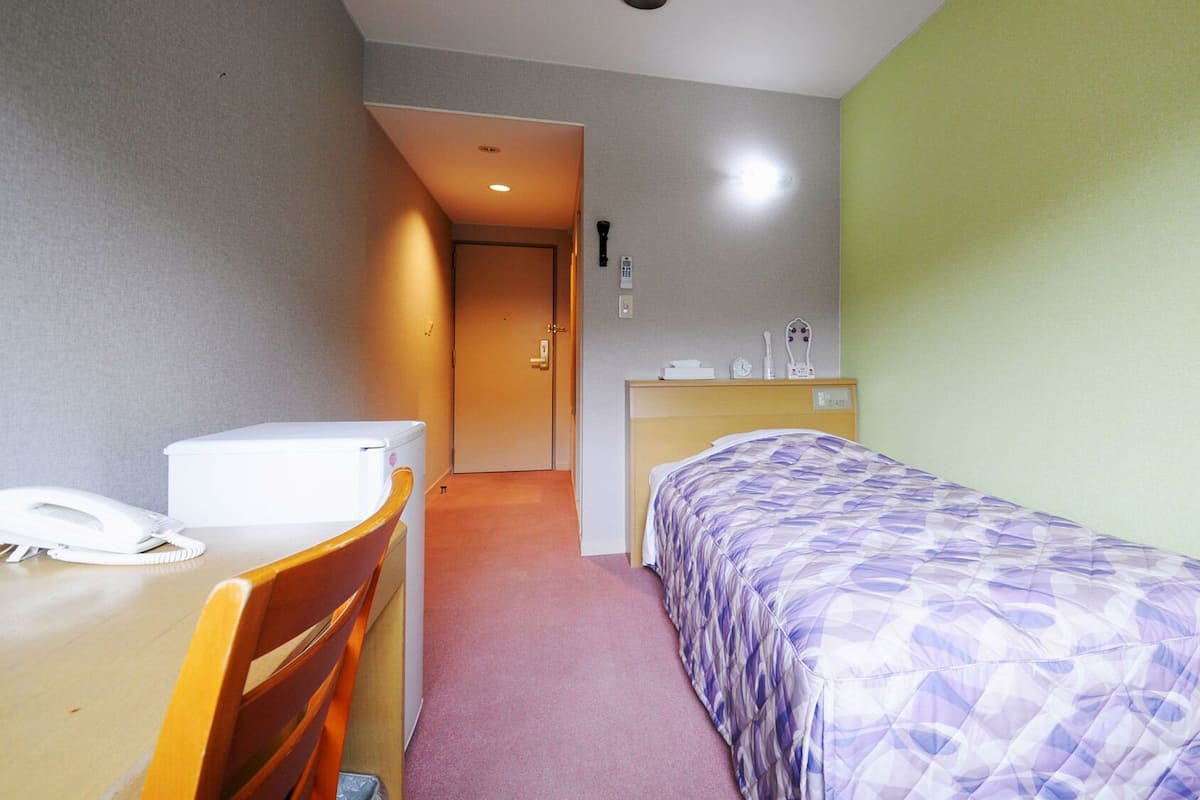 A hotel room with a bed, a desk with a telephone, a chair, a bedside table, and a door leading to another room.
