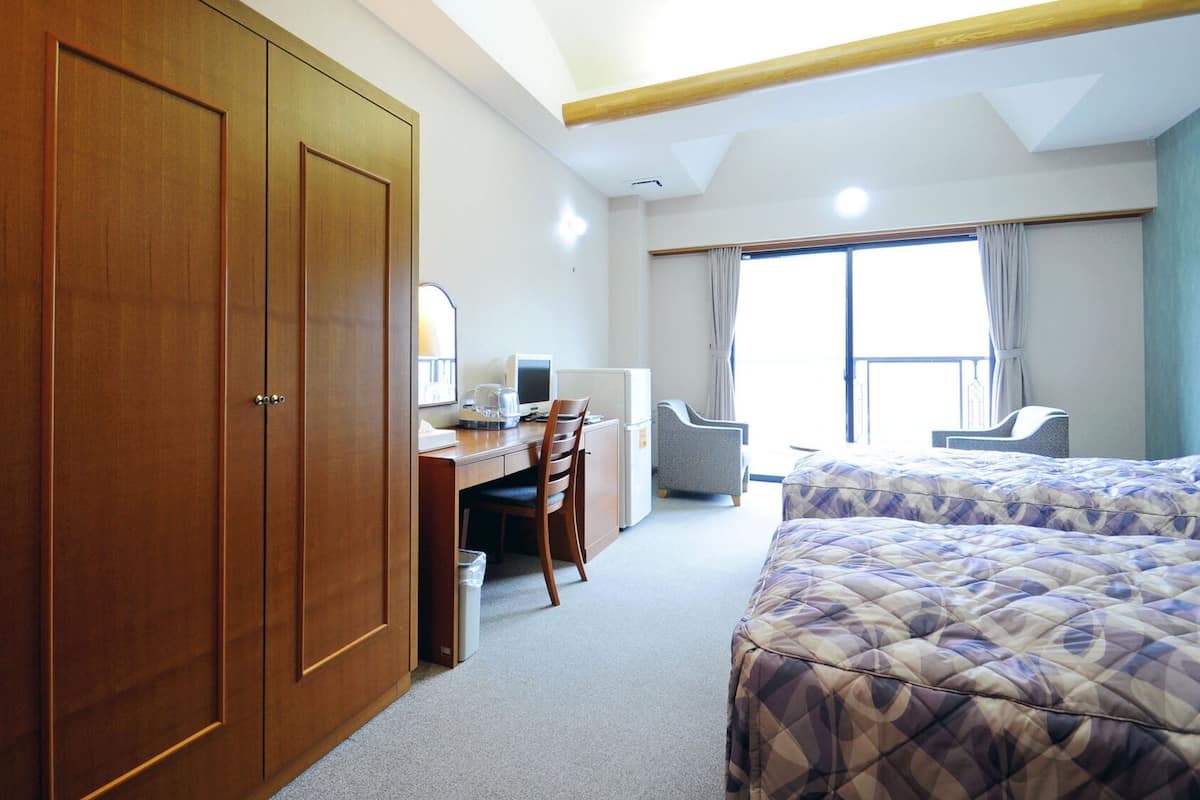A hotel room with a large wardrobe, a desk with a computer, a chair, a bed, and a window with curtains.