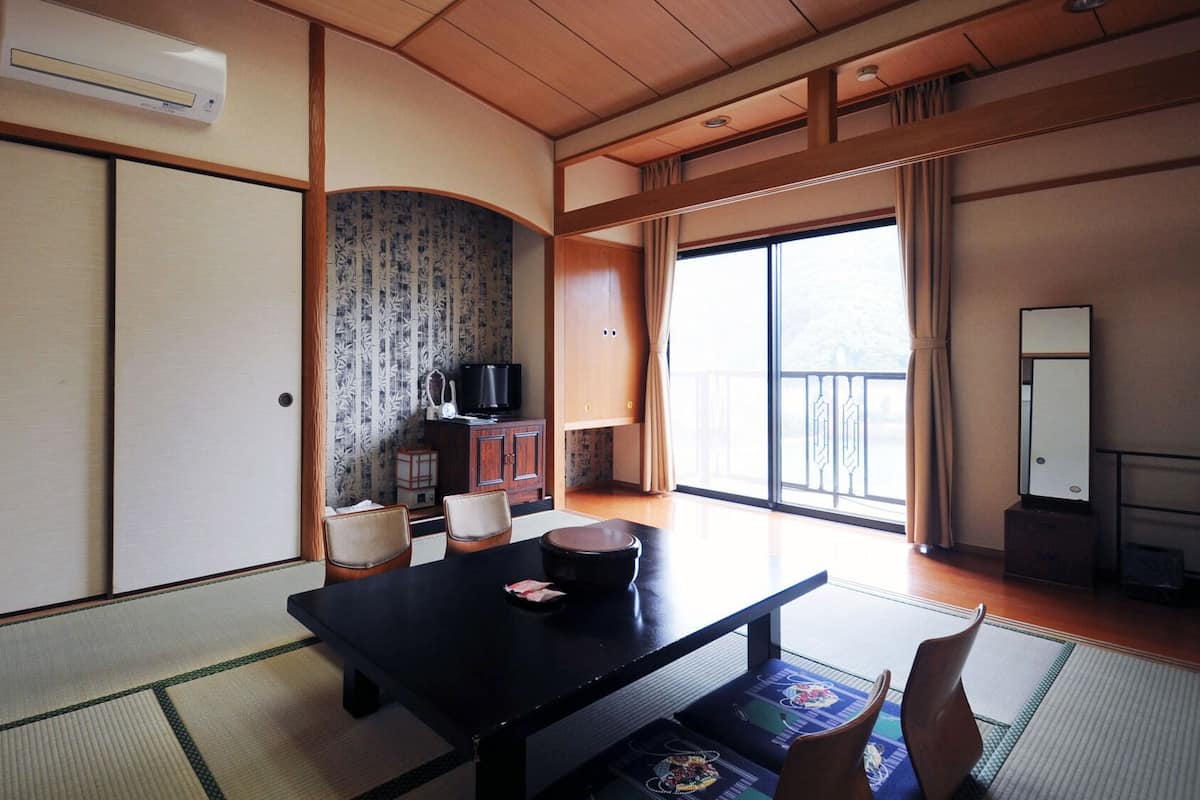 A traditional Japanese room with tatami flooring, a low table with cushions, a television on a cabinet, and a sliding door.