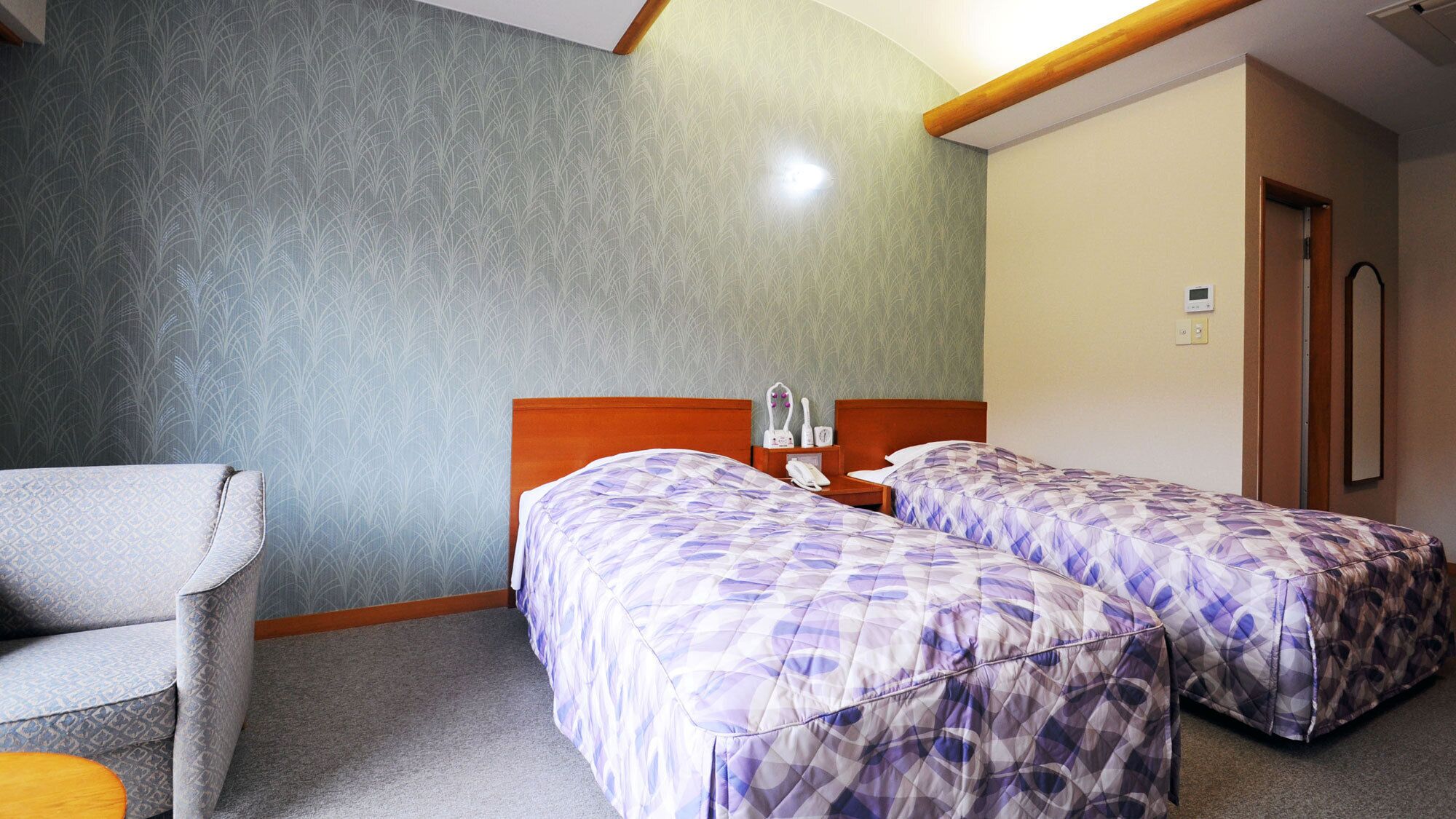 A hotel room with two beds, a chair, a bedside table, and a door.