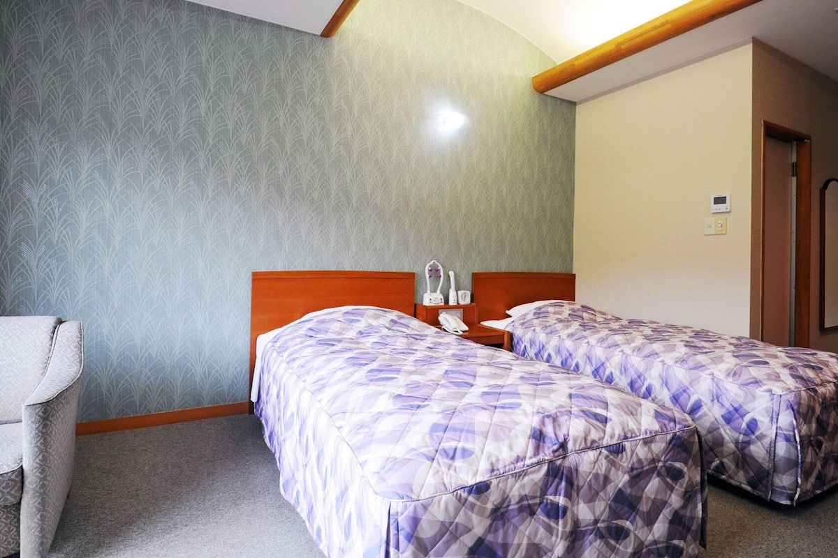 A hotel room with two beds, a chair, a bedside table, and a door.