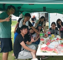 Un grupo de personas reunidas alrededor de una mesa con comida y bebidas, bajo una carpa verde.