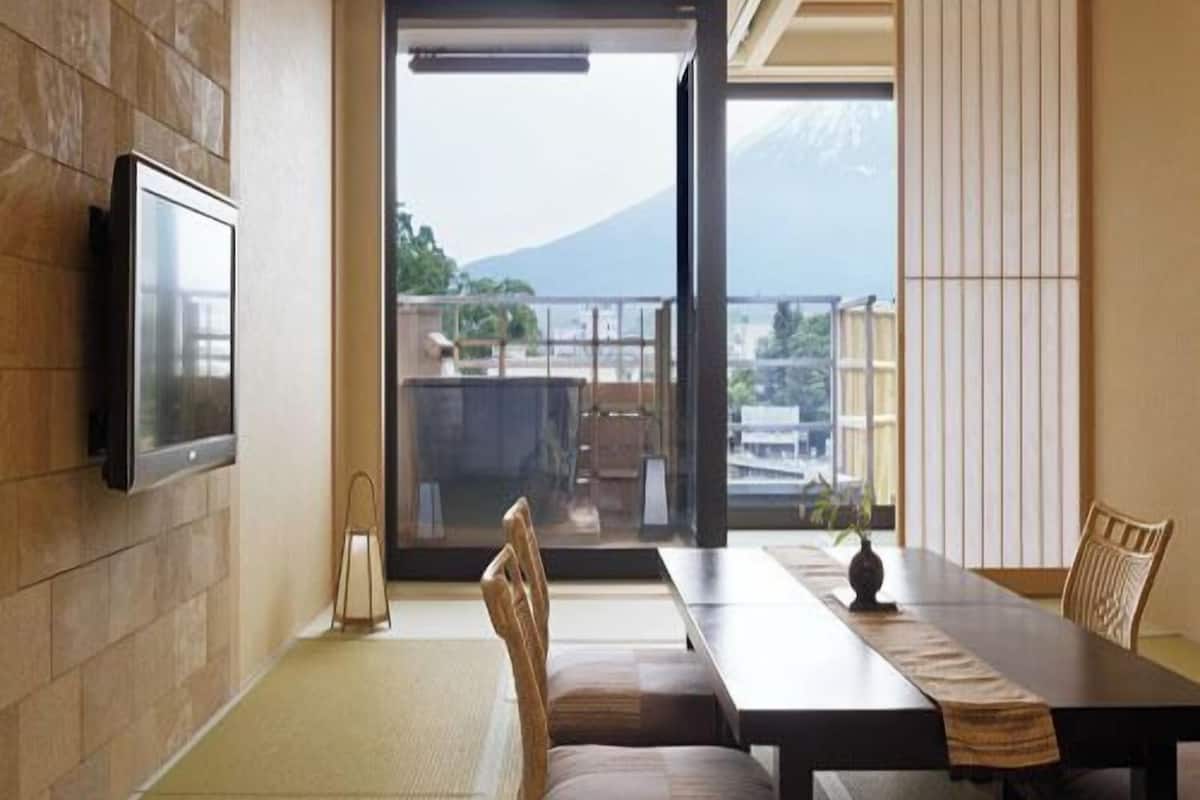 A room with tatami flooring, a low table, chairs, a television, and a sliding glass door leading to a balcony with a mountain view.