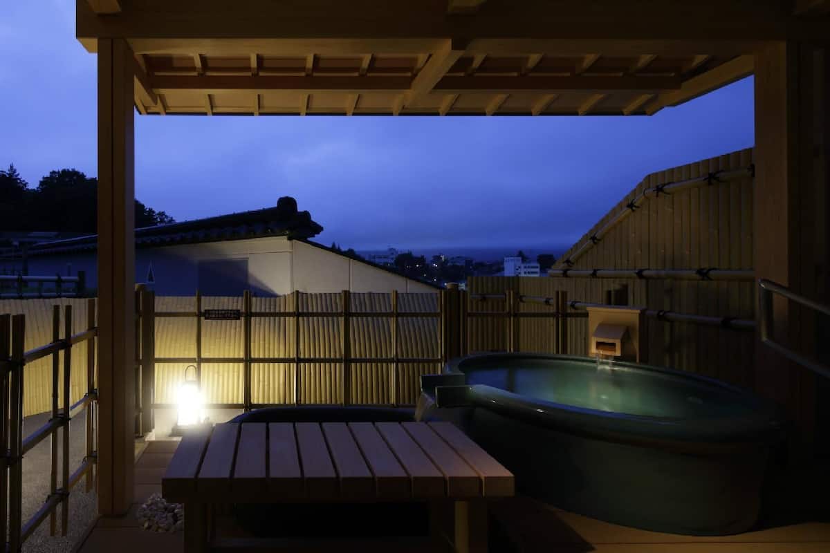 A wooden outdoor area with a hot tub, a wooden bench, and a view of a distant building.