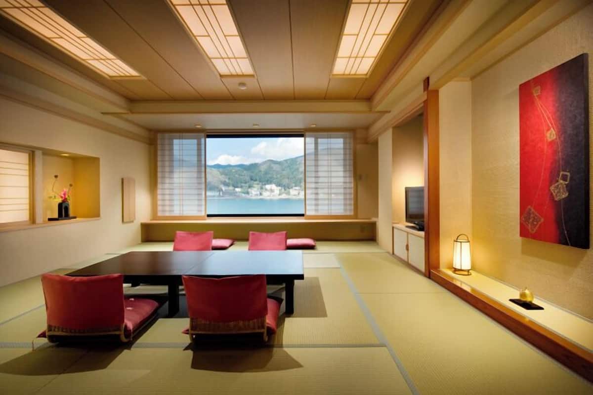 A traditional Japanese-style room with tatami flooring, a low table with cushions, a television, and a view of a mountainous landscape.
