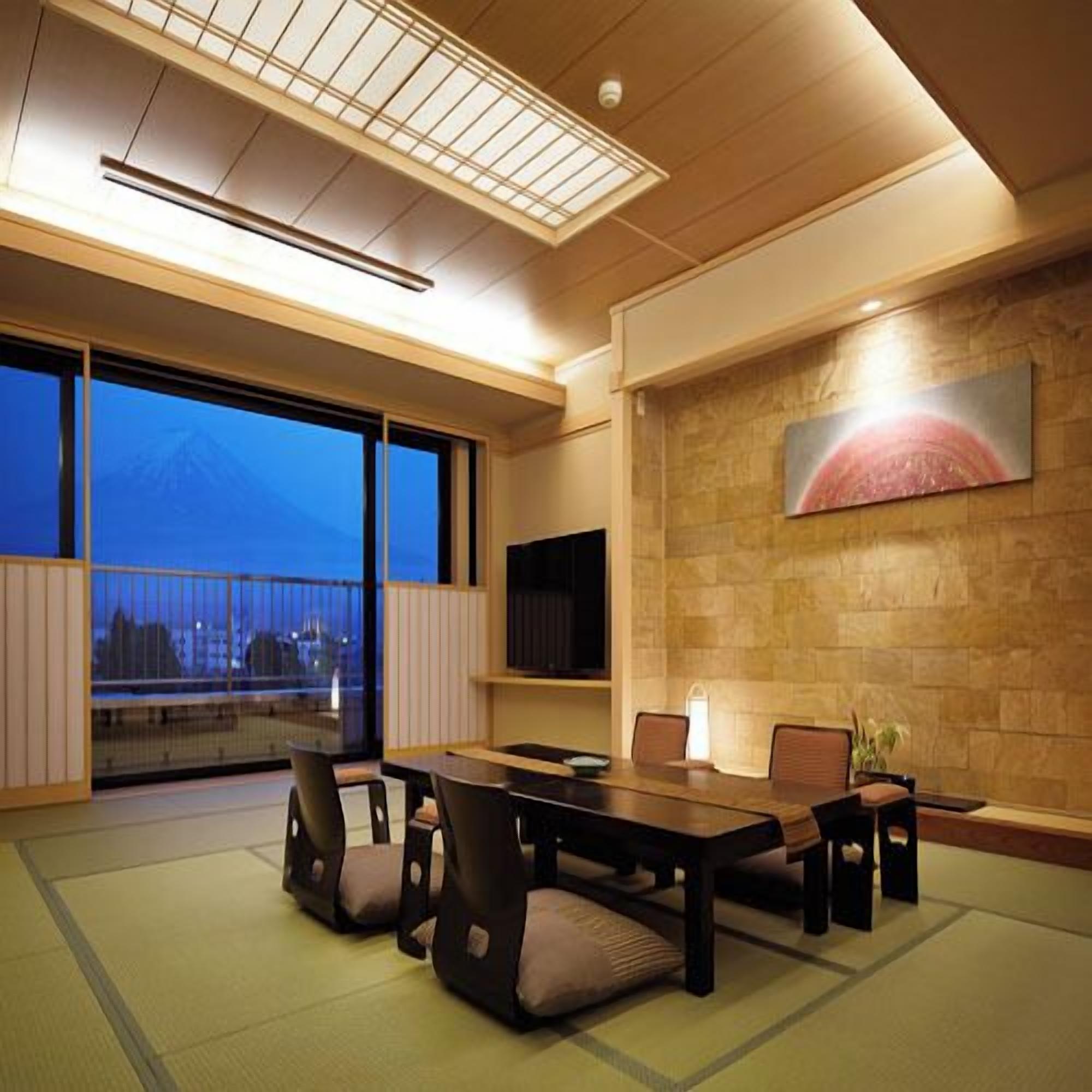 A modern interior with tatami flooring, a dining table, chairs, a mounted TV, and a large window with a view of a mountain.