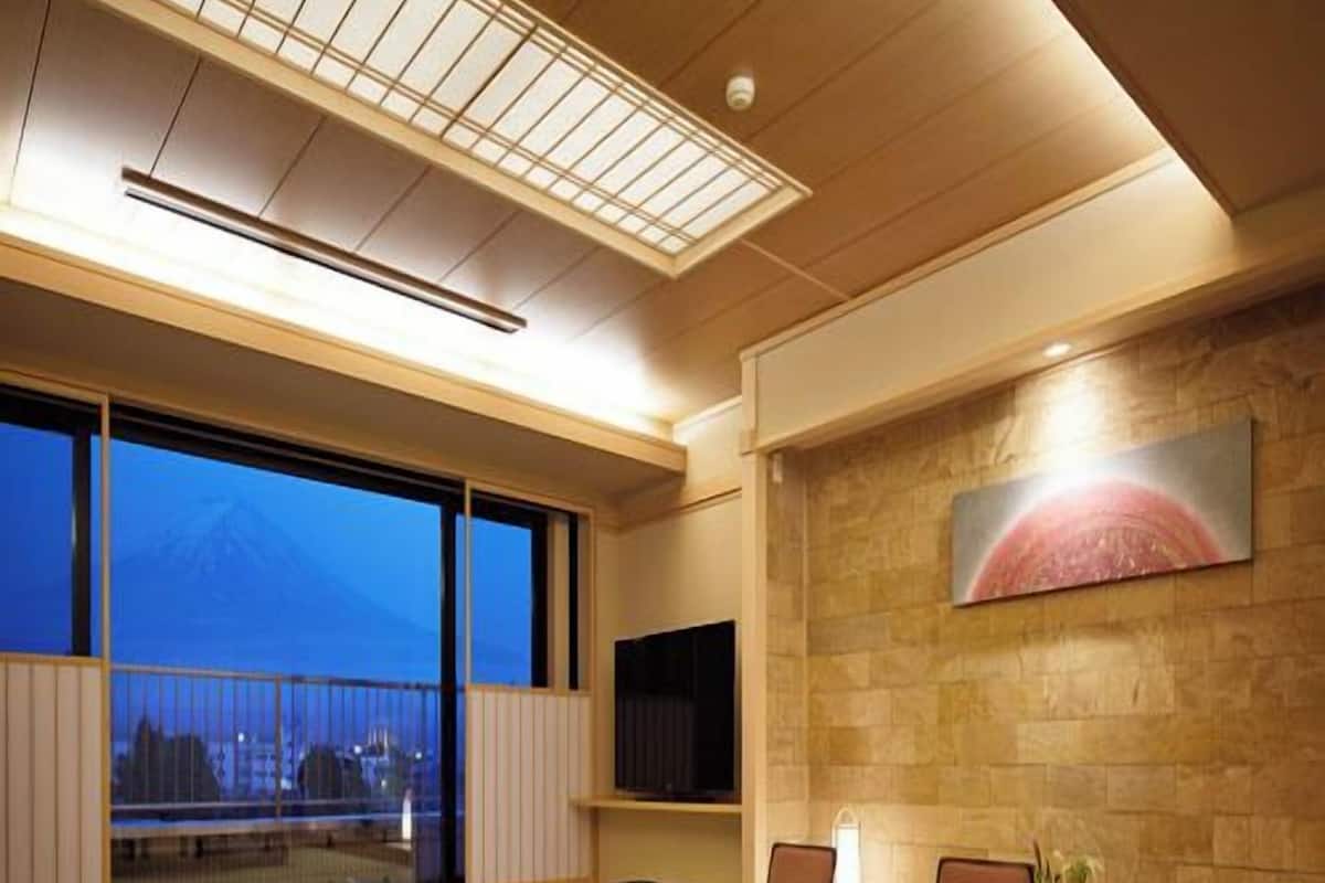 A modern interior with tatami flooring, a dining table, chairs, a mounted TV, and a large window with a view of a mountain.