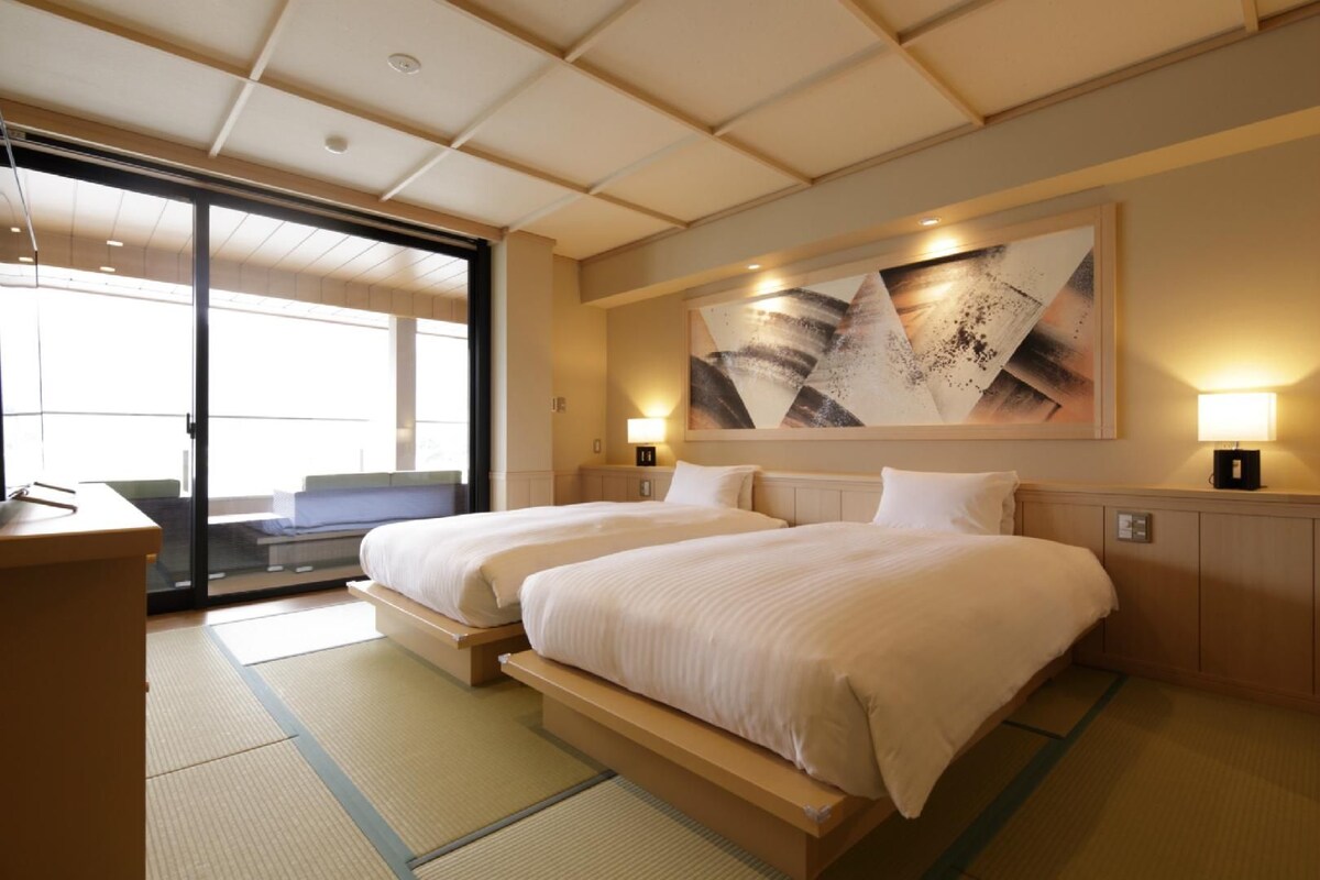Habitación de hotel con dos camas, un cuadro abstracto grande, puertas corredizas de vidrio y un escritorio.