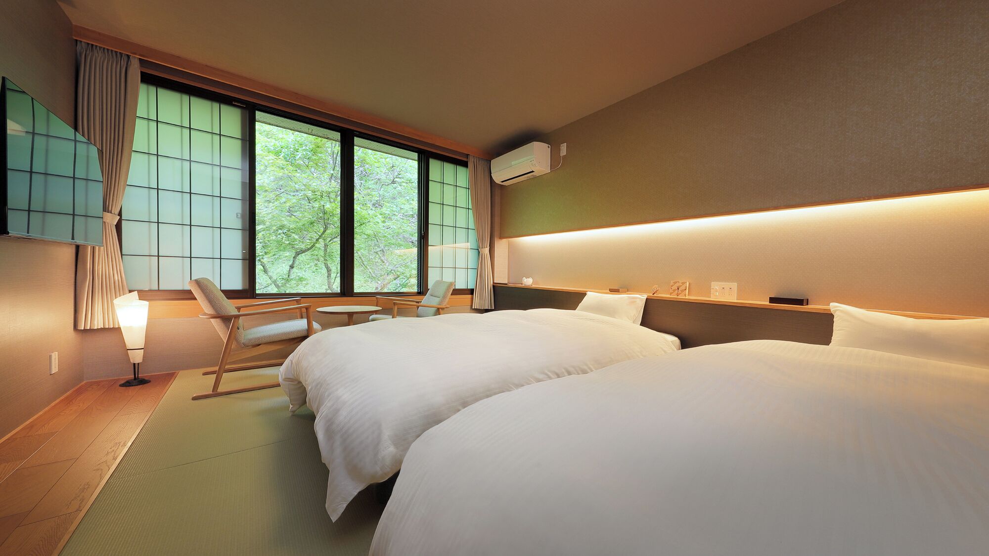 Ein Hotelzimmer mit zwei Betten, einer Sitzecke inklusive Sessel und Tisch, einem großen Fenster mit Vorhängen sowie einer Wand-Klimaanlage.