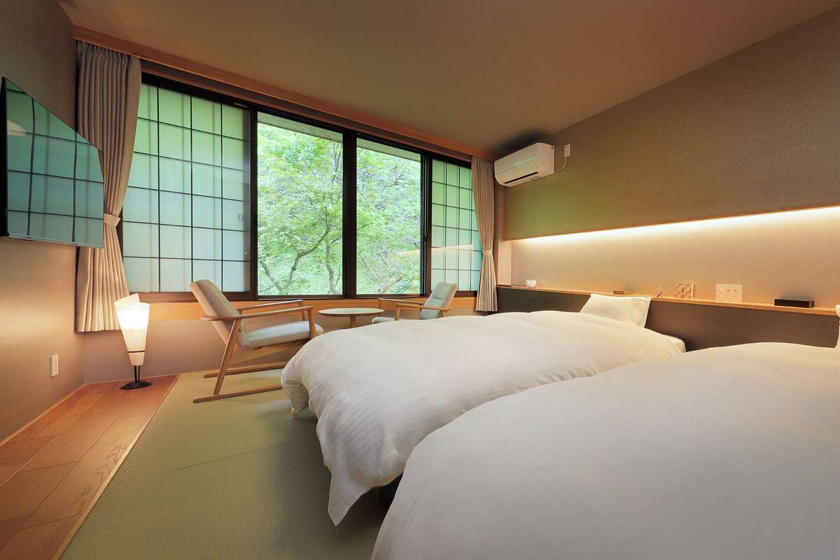 Une chambre d’hôtel avec deux lits, un coin salon comprenant une chaise et une table, une grande fenêtre avec des rideaux et un climatiseur fixé au mur.