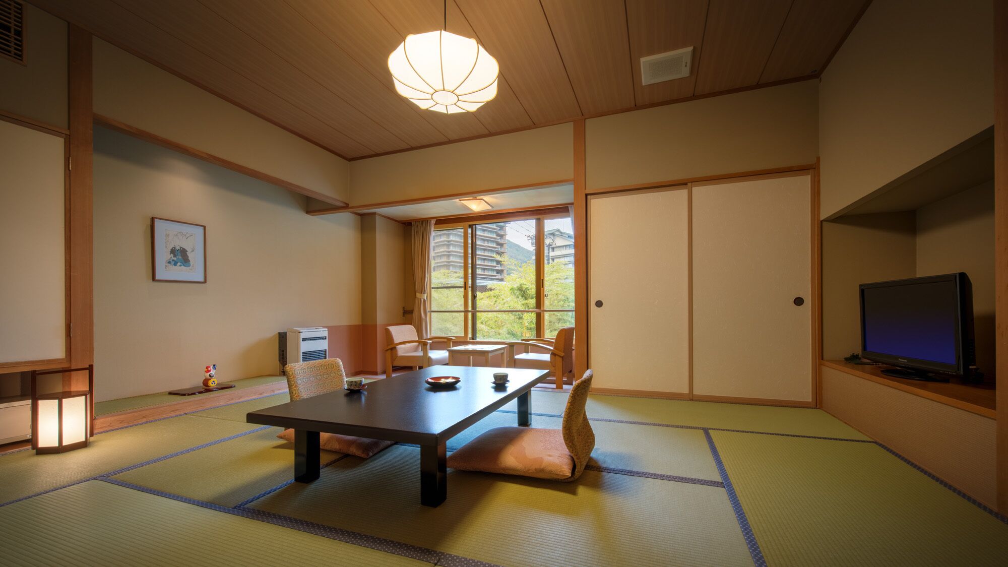 A traditional Japanese room with tatami flooring, a low table, chairs, a television, and a view of buildings outside.