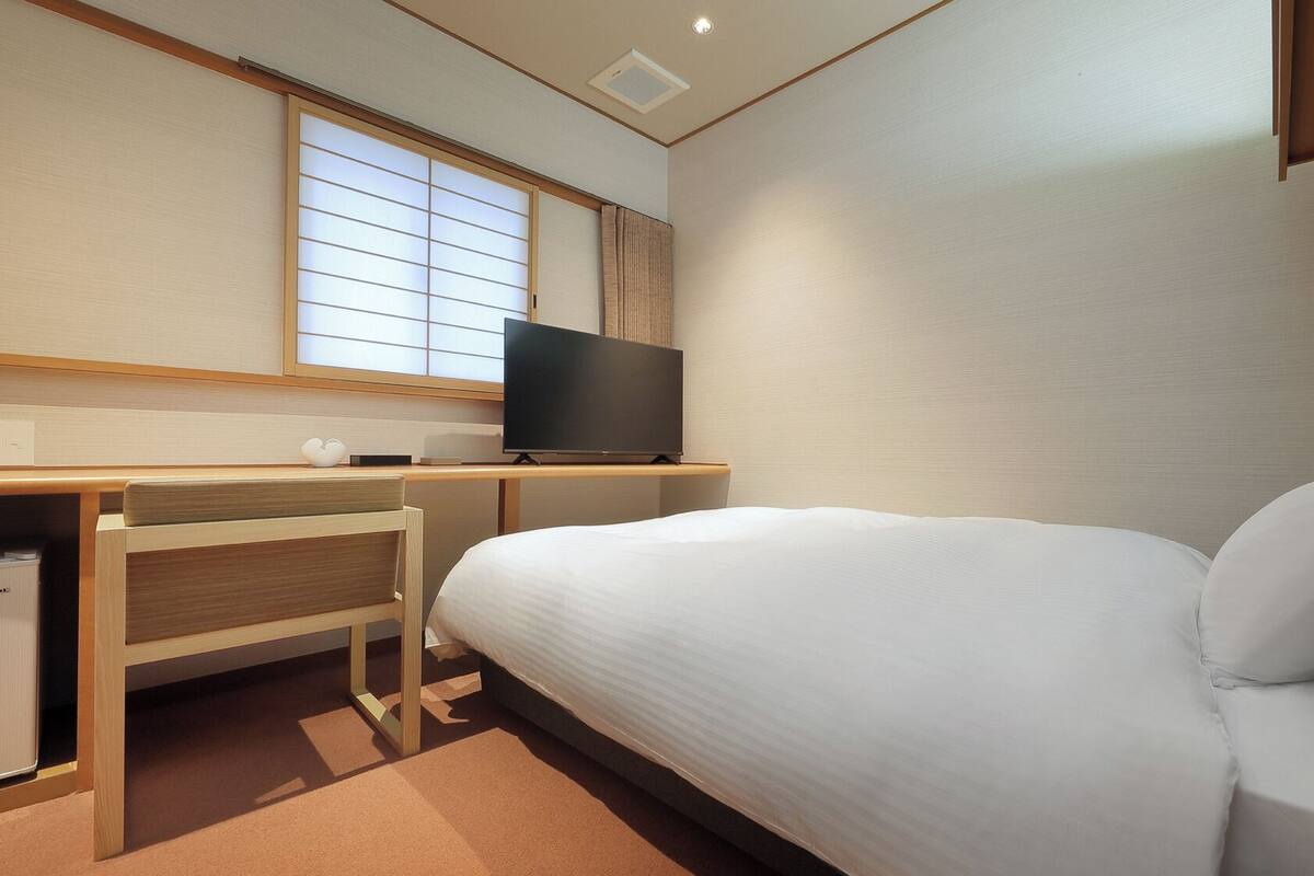 Une chambre d’hôtel avec un lit, un bureau avec une télévision et une fenêtre avec des stores.