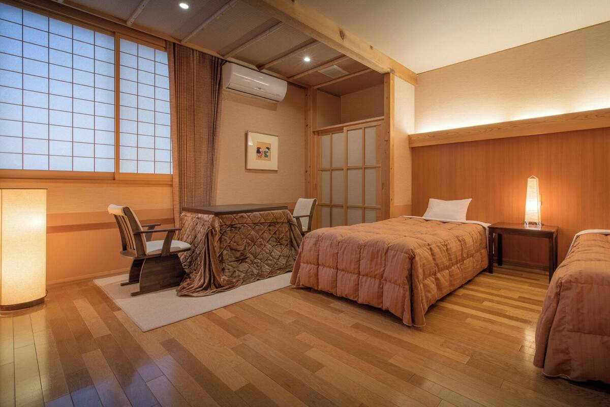 Une chambre d’hôtel avec un plancher en bois, deux lits, un bureau avec une chaise, une fenêtre avec des rideaux et un appareil de climatisation.