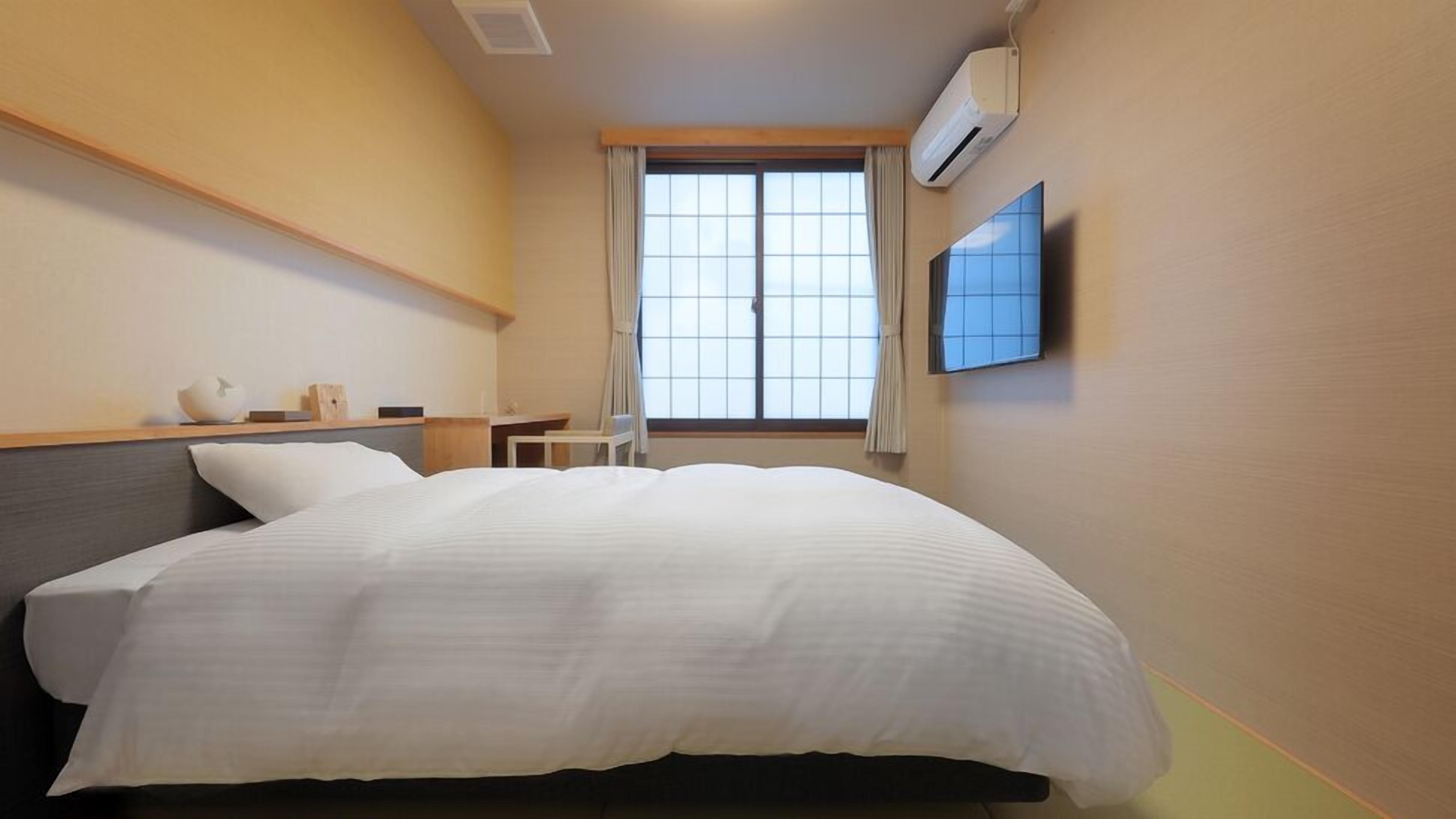 A hotel room with a bed, a small table, a chair, a TV, and a window with curtains.