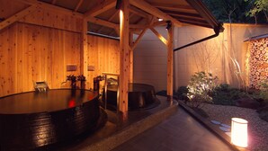 A wooden spa with hot tubs, a stone wall, and a stack of firewood.