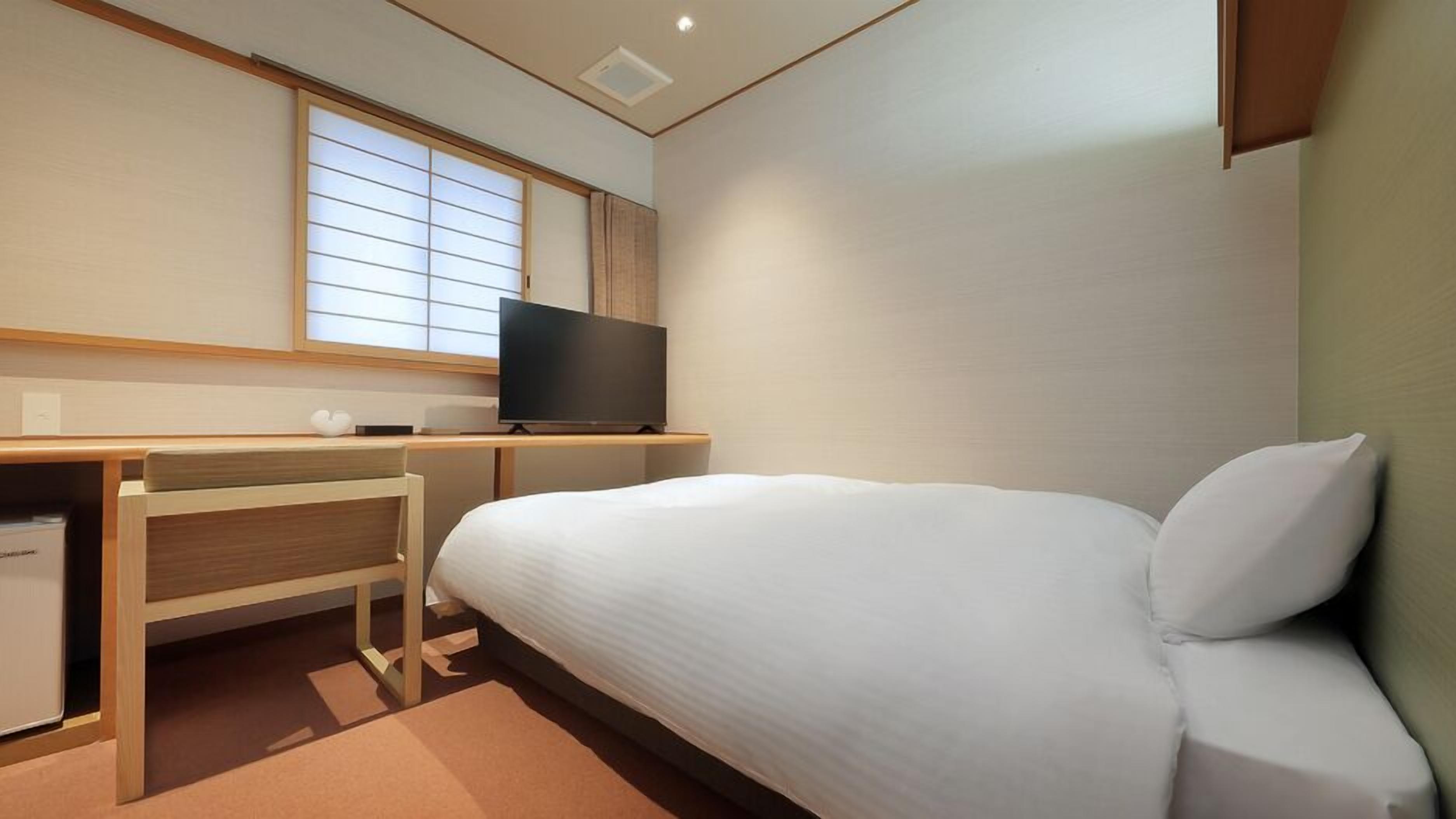 A hotel room with a bed, a desk with a television, and a window with blinds.