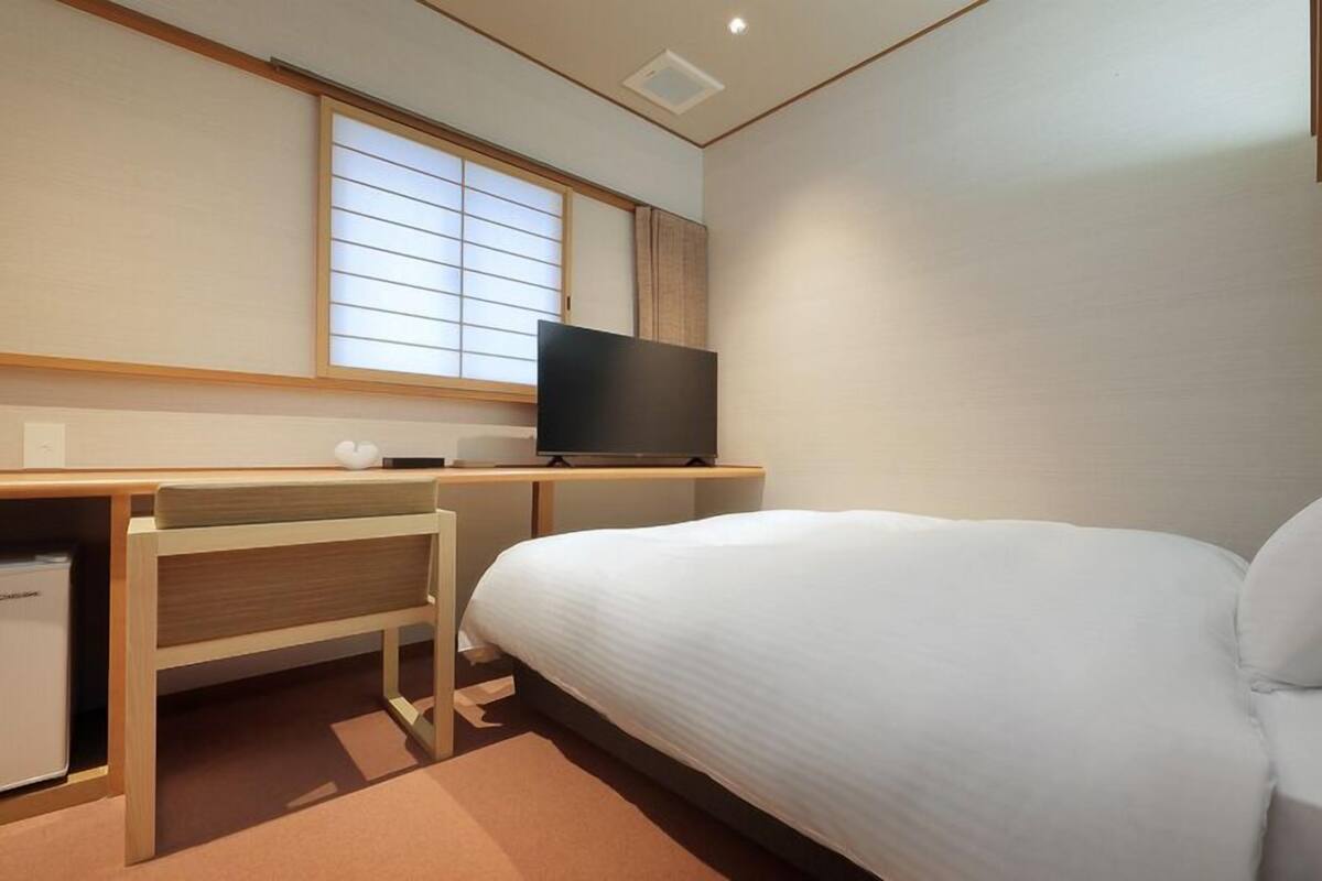 Une chambre d’hôtel avec un lit, un bureau avec une télévision et une fenêtre avec des stores.