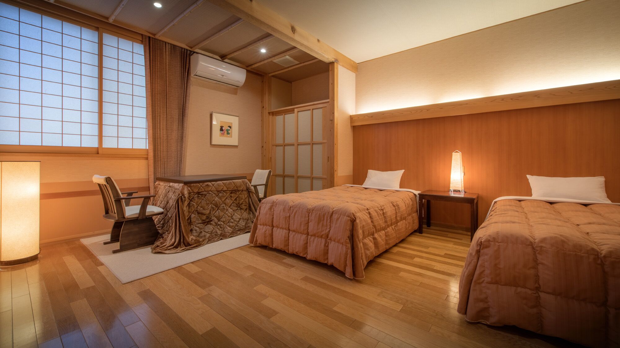 A hotel room with a wooden floor, two beds, a desk with a chair, a window with curtains, and an air conditioning unit.