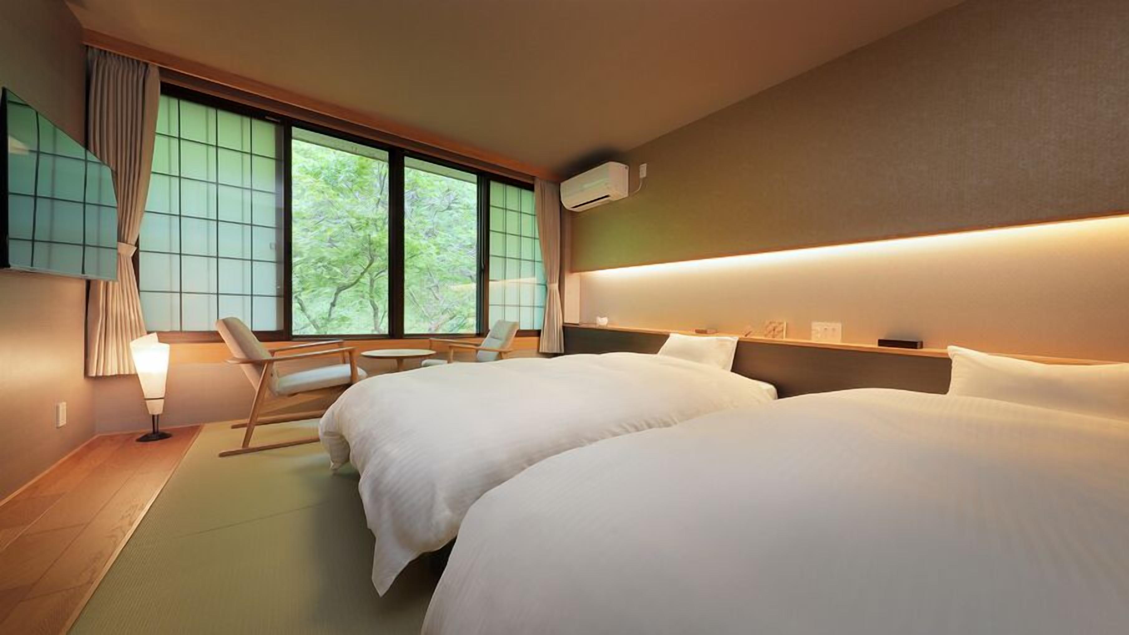 A hotel room with two beds, a sitting area with a chair and table, and a large window with curtains.