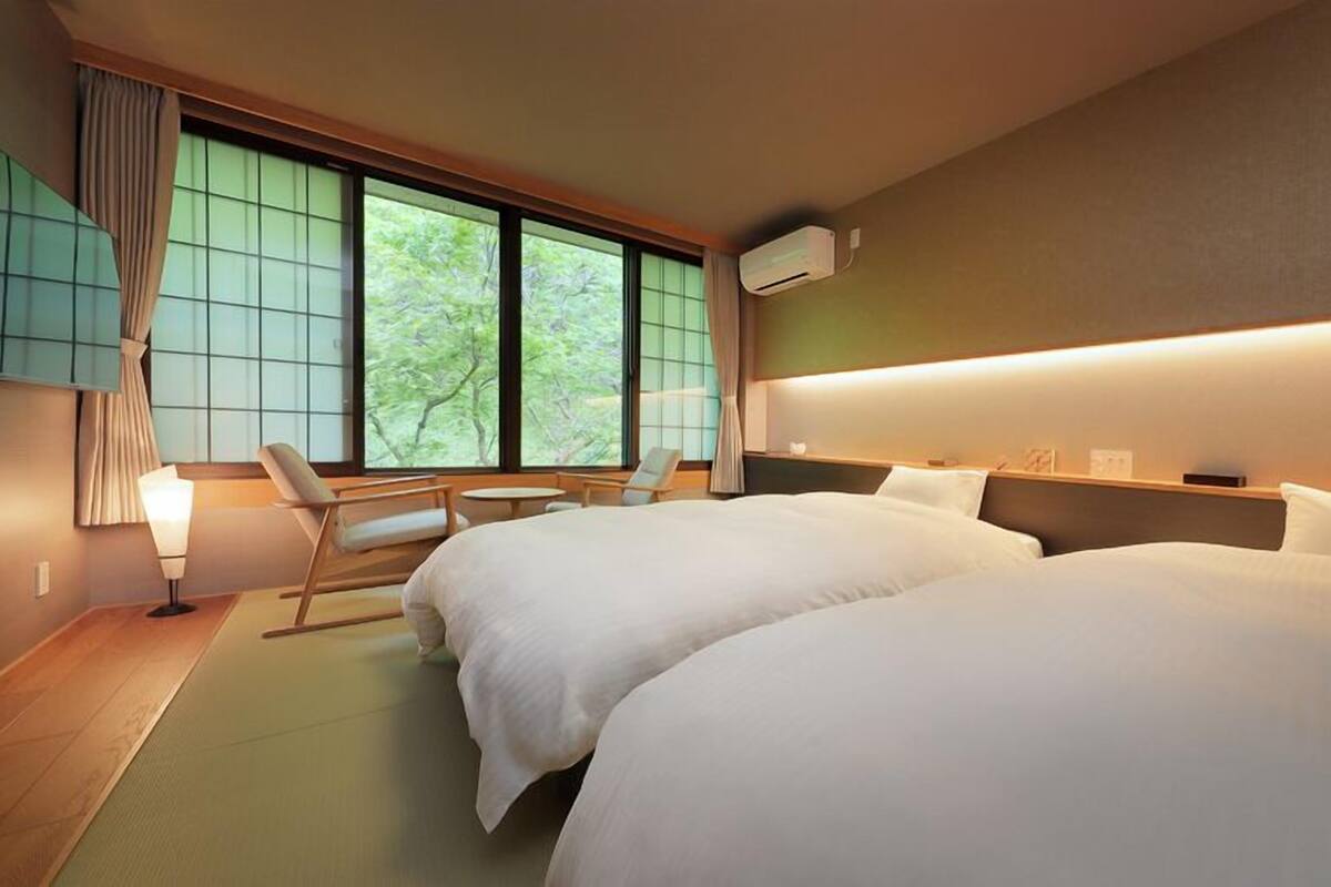 Une chambre d’hôtel avec deux lits, un coin salon avec une chaise et une table, et une grande fenêtre avec des rideaux.