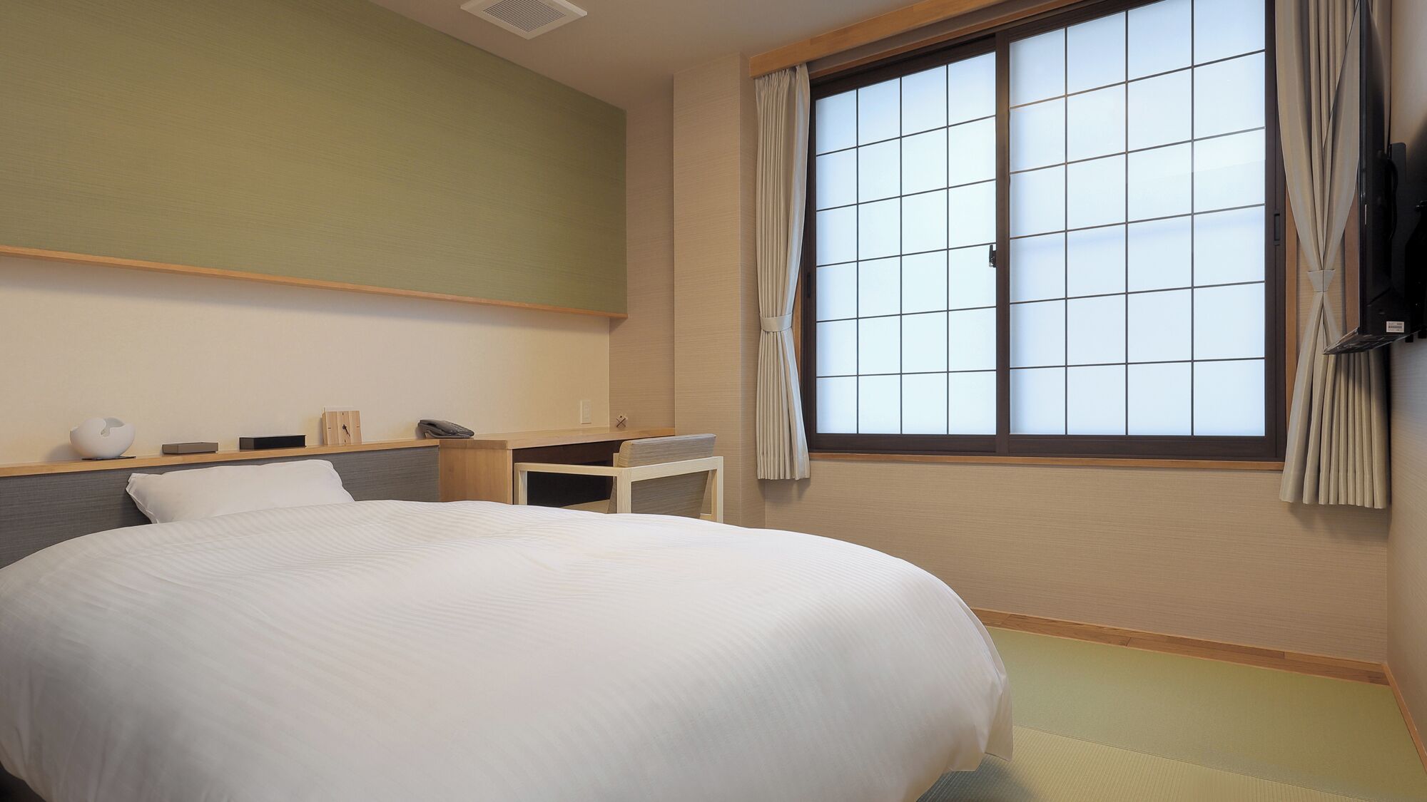 Ein Hotelzimmer mit einem großen Bett, einem kleinen Schreibtisch, einem Stuhl, einem Fenster mit Vorhängen und einem an der Wand befestigten Regal.
