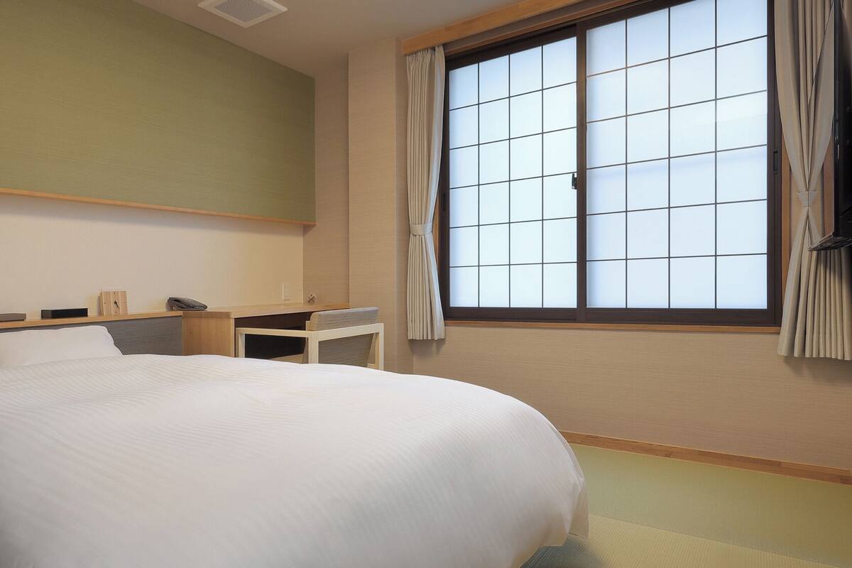 Une chambre d’hôtel avec un grand lit, un petit bureau, une chaise, une fenêtre avec des rideaux et une étagère fixée au mur.