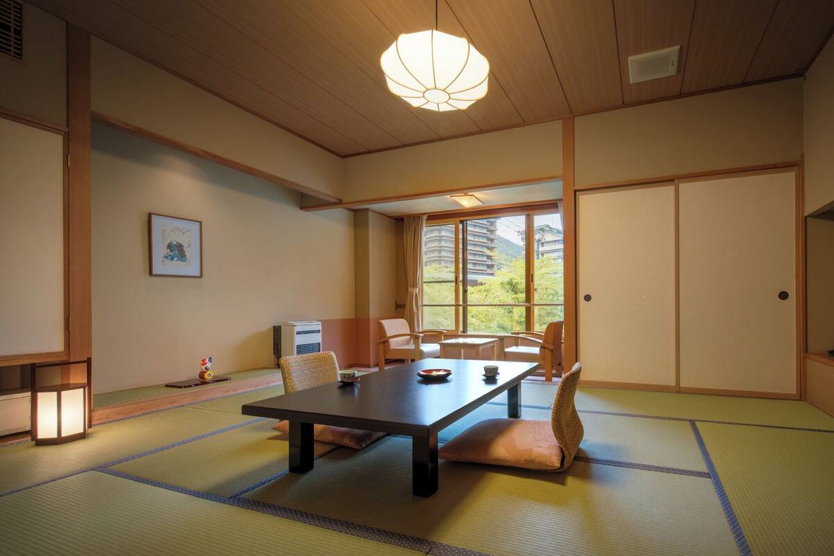 Une pièce de style japonais traditionnel, avec un sol recouvert de tatamis, une table basse, des chaises, un téléviseur et une vue sur les bâtiments à travers de grandes fenêtres.