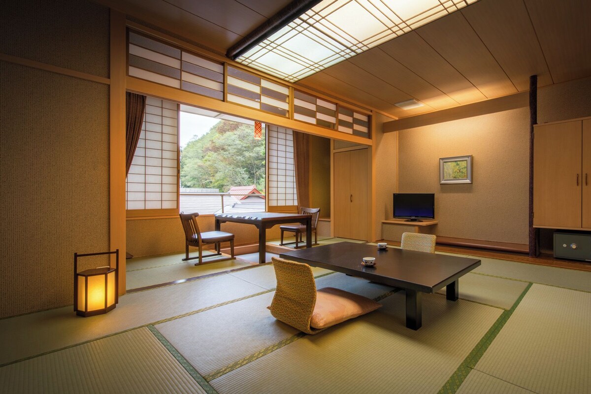 Una stanza tradizionale giapponese con pavimento in tatami, un tavolino, sedie, un televisore e una porta scorrevole.