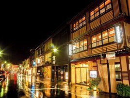 Rua tradicional japonesa à noite, com edifícios iluminados e pavimento molhado refletindo luzes.