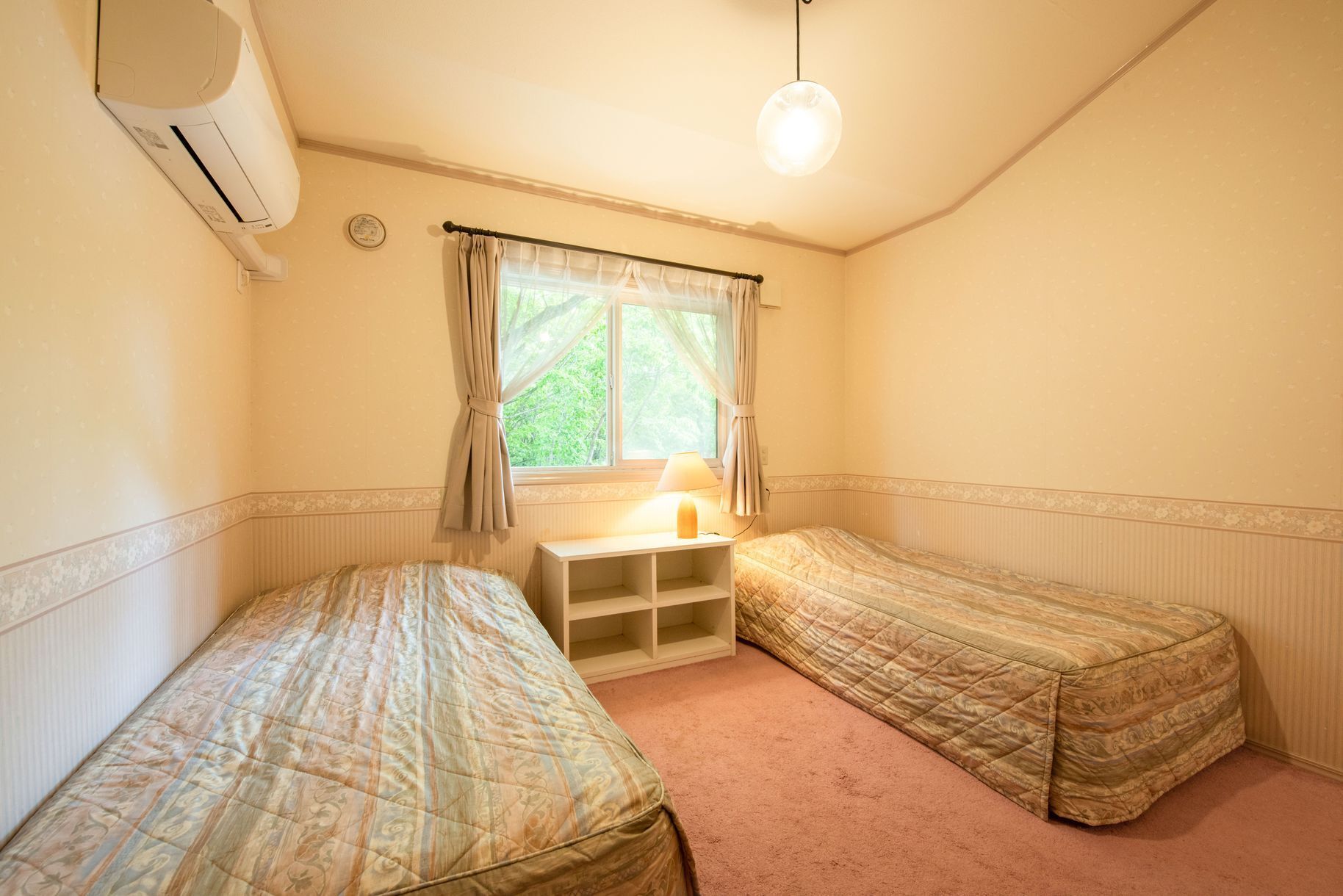 A room with two single beds, a bedside table with a lamp, a window with curtains, and an air conditioning unit.
