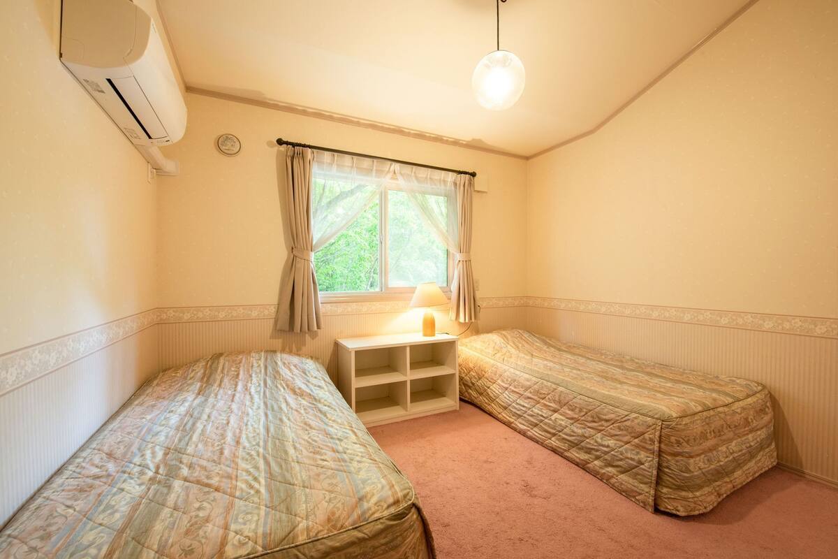Ein Zimmer mit zwei Einzelbetten, einem Nachttisch mit Lampe, einem Fenster mit Vorhängen und einer Klimaanlage.