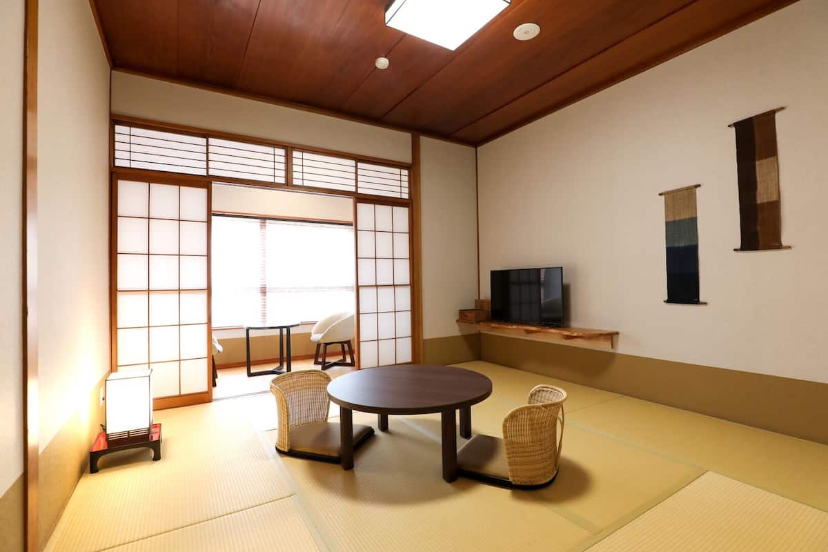 A traditional Japanese room with tatami flooring, a tatami mat table, two low chairs, a television on a shelf, and sliding shoji doors.