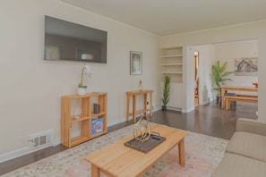 Uma sala de estar com televisão, estantes de madeira, uma mesa de centro e um espaço para refeições.