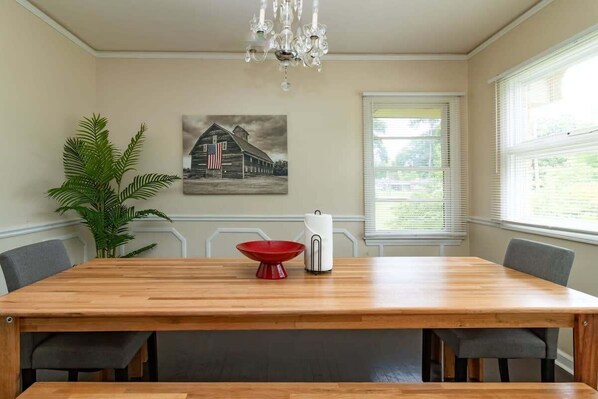 Uma área de jantar com mesa de madeira, cadeiras, uma tigela vermelha, um eletrodoméstico branco, um lustre, uma planta e uma pintura de um celeiro.