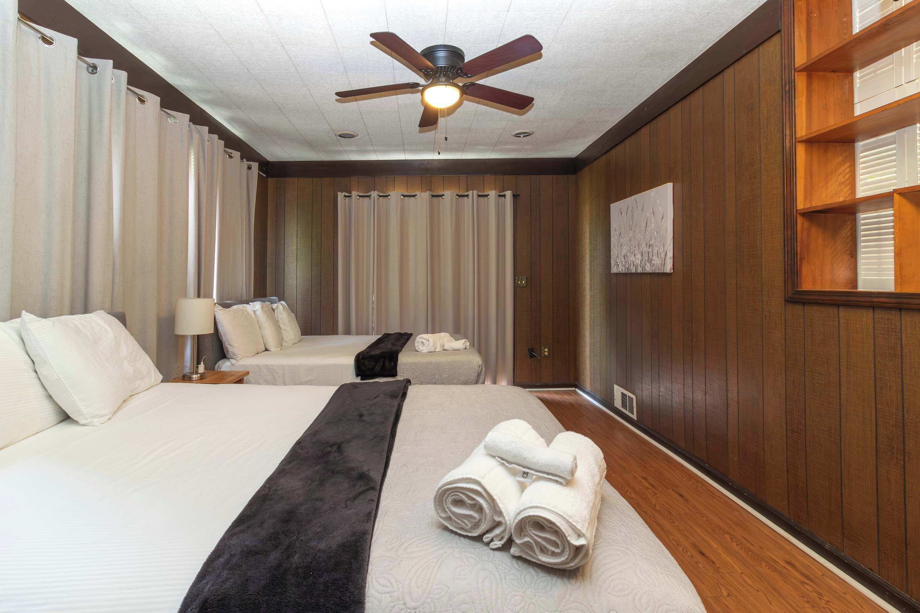 A hotel room with two beds, a ceiling fan, a wooden wall, and a painting.
