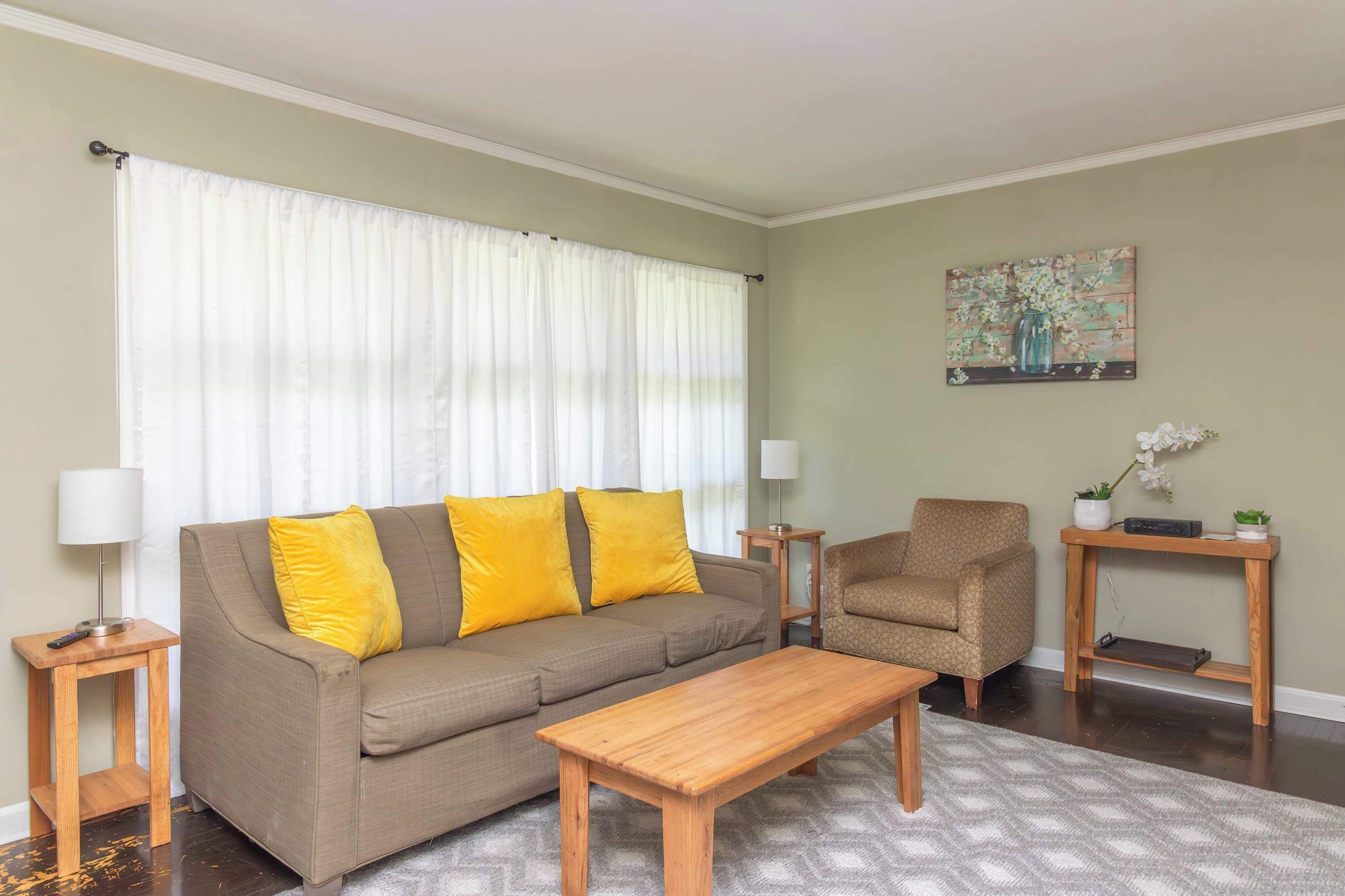 A living room with a sofa, a wooden coffee table, a side table with a lamp, a chair, and a small wooden side table with a plant.