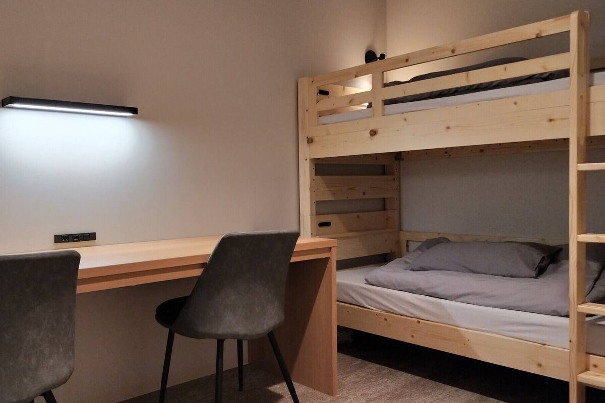 Uma cama beliche com escrivaninha, duas cadeiras, mesa de cabeceira e luminária fixa na parede.