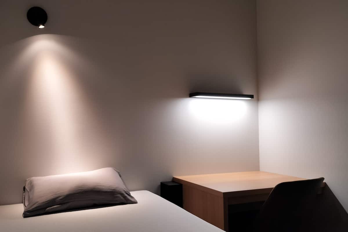 Quarto pequeno e minimalista com uma cama, uma escrivaninha e uma luminária na parede.