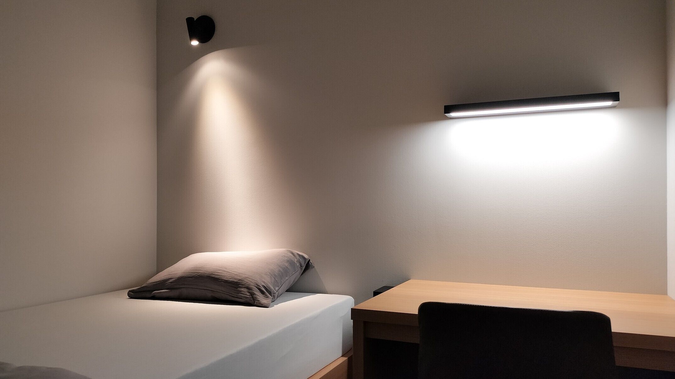 Un dormitorio con cama, escritorio y lámpara de pared.